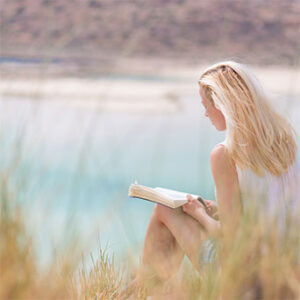 Woman sitting by a calm lake reading a book on a warm, sunny day.