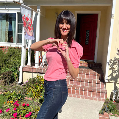 Gretchen Hydo standing outside on a warm, sunny day, using her hands to make the shape of a heart.