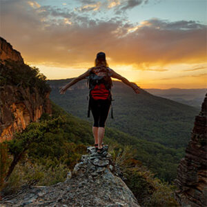 Woman walking on narrow mountain path toward orange sunset, looking ahead over forest-covered valley.