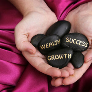 Woman's hands resting on red satin cloth, holding black stones with the following words painted in gold lettering on them: Wealth, Growth, Success